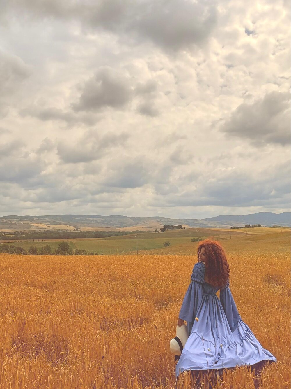 Kobieta w błękitnej sukni flying dress spacerująca po polu pszenicy pod pochmurnym niebem